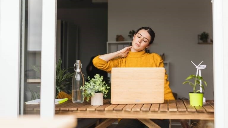 Eine Frau sitzt am Schreibtisch und berührt mit ihrer Hand ihren Nacken