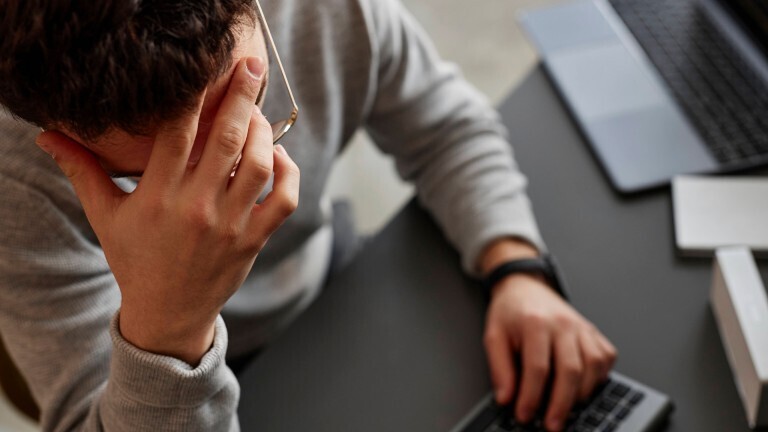 Ein Mann sitzt vor einem Computer und einem Laptop an einem Schreibtisch vor. Wir sehen ihn von oben. Er hält seine Hand gegen seine Stirn.
