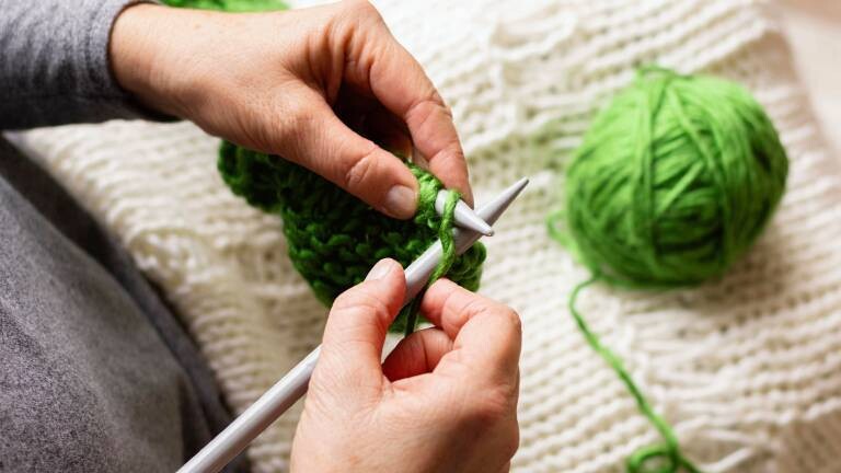 Eine Frau strickt mit einem dicken, grünen Wollfaden und zwei dicken Sticknadeln. Im Hintergrund liegt ein bereits mit dicker, weißer Wolle gestricktes Stück.
