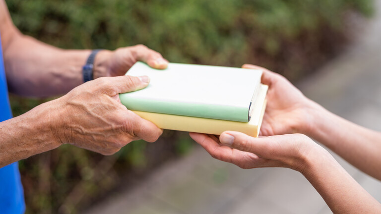 Hände einer männlichen Person übergeben zwei Bücher in die Hände einer weiblichen Person.