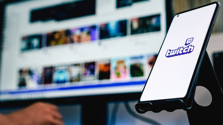 Auf einem Computer-Bildschirm ist die Startseite der Livestream-Plattform Twitch geöffnet. Daneben ist ein Handy in einer Halterung aufgestellt, auf der das Twitch-Logo zu sehen ist.