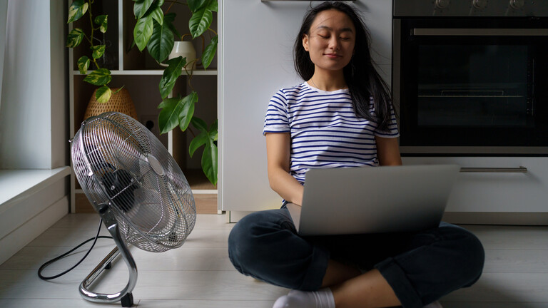 Eine Freiberuflerin sitzt im Homeoffice mit einem Laptop auf dem Boden und wird von einem elektrischen Ventilator angeweht.