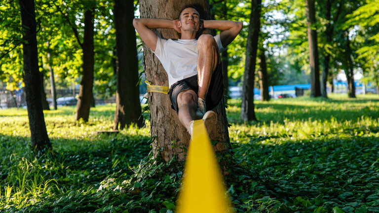 Ein Mann sitzt an einem Baum gelehnt auf einer Slackline und ruht sich zufrieden aus.