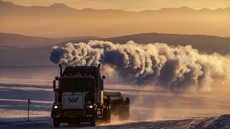 LKW auf verschneiter Piste mit großer Abgaswolke vor Bergen im Winter, Abendlicht, Dalton Highway, Alaska