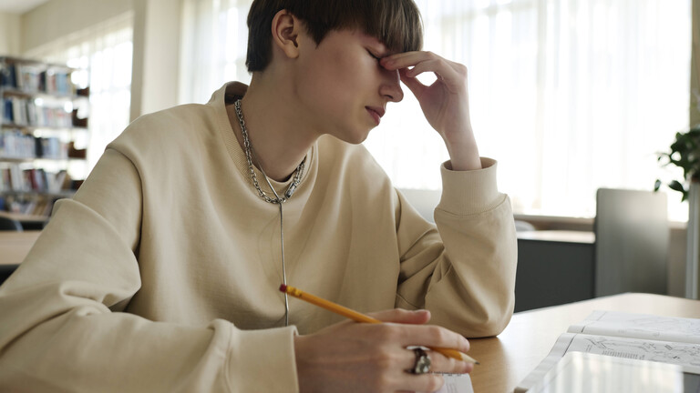 Eine Person sitzt in einer Bibliothek an einem Schreibtisch und hält einen Stift in der Hand. Mit der anderen Hand hält sich die Person an der Nase fest. Die Augen sind geschlossen.
