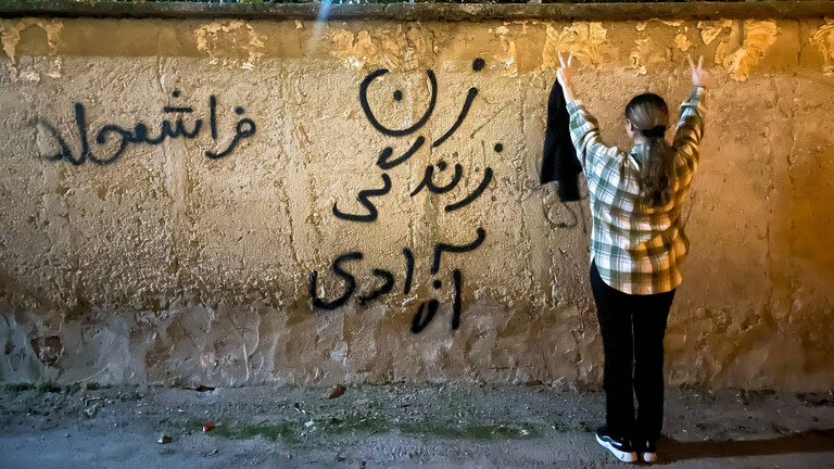 Eine junge Frau im Iran steht – ohne den obligatorischen Hijab – vor einer Mauer, mit dem Schriftzug: Frau, Leben, Freiheit.