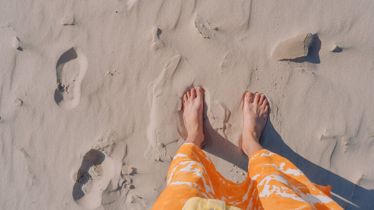 Eine Frau steht in einem orangefarbenen Rock barfuß am Strand.