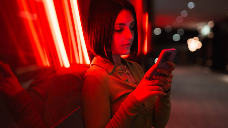 Eine Frau lehnt an die Glasfassade eines Gebäudes und schaut auf ihr Handy. Hinter dem Glas sind rote Neonröhren eingebaut, die die Frau und das Gebäude in rotes Licht umhüllen.