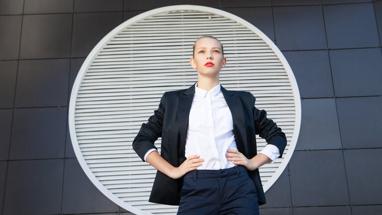 Eine Business-Frau in schwarzem Hosenanzug und mit weißer Bluse hat die Hände in die Hüften gestemmt und schaut in die Ferne.