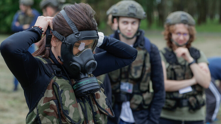 Eine junge Frau probiert eine Gasmaske an. Im Hintergrund stehen junge Menschen in Flecktarn, die sie ansehen