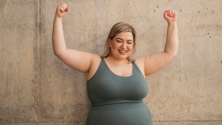 Eine Frau hebt ihre Arme hoch. Sie trägt ein Sport-Outfit und steht vor einer leeren Betonwand.