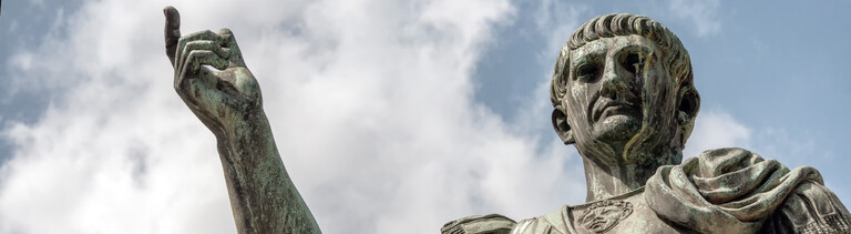 Untersichtige Fotografie einer Bronzestatue des römischen Kaisers Caesar Nervae Trajan vor weiß bewölktem blauen Himmel