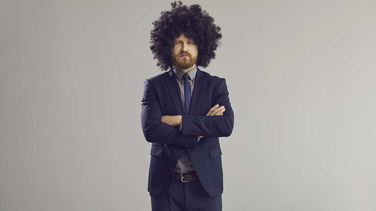 Ein Mann in Anzug und mit einer Perücke steht mit verschränkten Armen vor grauem HIntergrund und blickt in die Kamera.