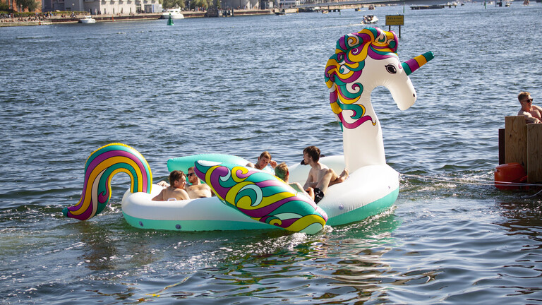 Eine Gruppe junger Leute, die in einem riesigen aufblasbaren Boot in Form eines bunten Cartoon-Einhorns im Wasser eines städtischen Hafens in Kopenhagen in Dänemark Spaß haben.