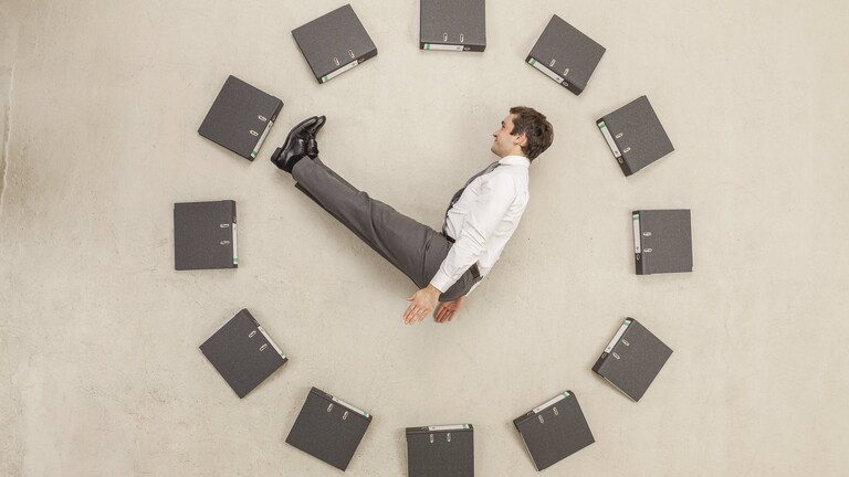 Aktenordner - kreisförmig auf dem Boden angeordnet - bilden eine Uhr. Ein Mann in Anzug stellt die Zeiger dar.