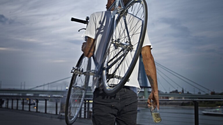 Ein Mann trägt sein Fahrrad auf der Schulter und hält eine Flasche in der rechten Hand.