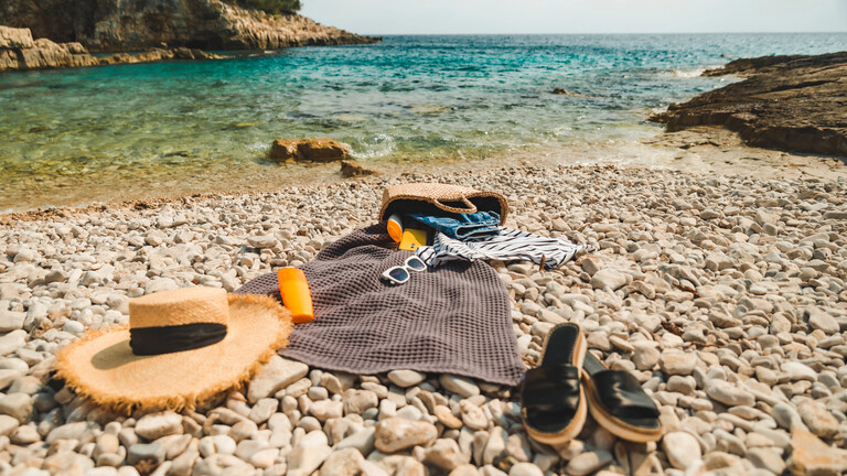Handtuch, Sonnenhut Latschen, Sonnenbrille und -milch liegen auf einem Steinstrand am Meer