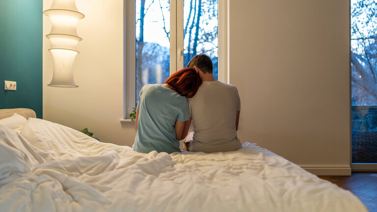 Eine Frau und ein Mann sitzen nebeneinander am Bettrand. Sie lehnt ihren Kopf an seiner Schulter an.
