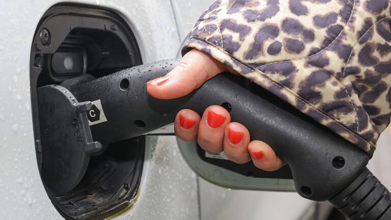 Foto einer jungen Frauenhand mit rot lackierten Nägeln, die aus einer Jacke mit Leopardenmuster ragt, die bei Regen ein schwarzes Ladekabel in ein weißes Auto steckt.