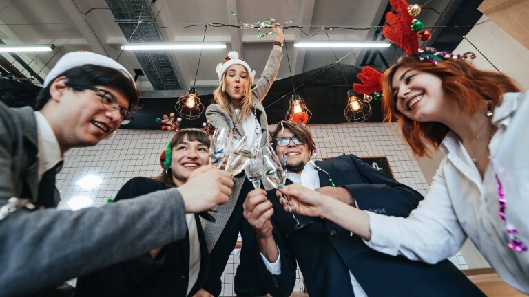 Eine Gruppe junger Arbeitskolleginnen und Arbeitskollegen stößt mit Sektgläsern gemeinsam an. Sie tragen dabei weihnachtliche Accessoires wie Weihnachtsmützen und Haarreifen mit Rentier-Geweihen aus Stoff.