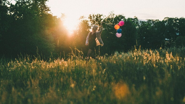 Eine Frau läuft auf einer Wiese. Hinter sich her zieht sie bunte Ballons. Die Sonne scheint. Es ist Abend.