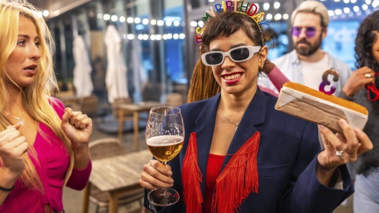 Eine Frau hält einen Kuchen in der Hand, in dem eine Kerze mit der Nummer drei steckt. In der anderen Hand hält sie ein Glas mit Bier. Sie trägt eine Sonnenbrille und einen Haarreif mit dem Schriftzug "It's my birthday".