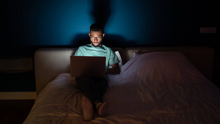Junger Mann arbeitet an einem Laptop, liegt nachts im Bett und schaut aufmerksam auf den Bildschirm.