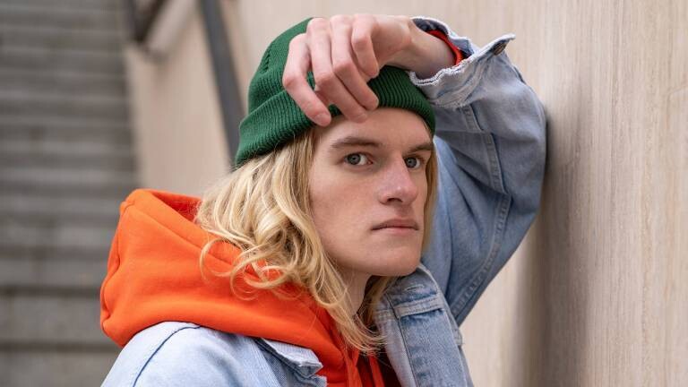 Frustriert schauender Mann mit grüner Mütze, orangen Hoodie und Jeansjacke lehnt an Wand