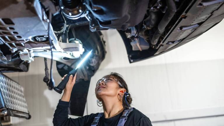 Mechanikerin inspiziert mit Taschenlampe die Unterseite eines Autos auf einer Hebebühne