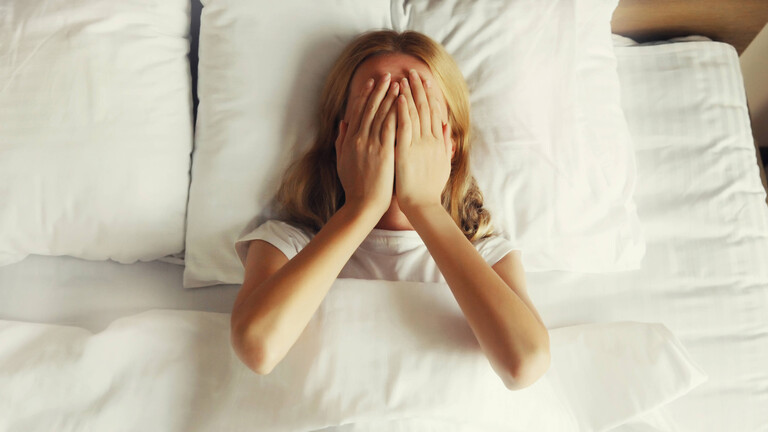 Frau, die auf dem Bett liegt und sich die Hände vor das Gesicht hält.