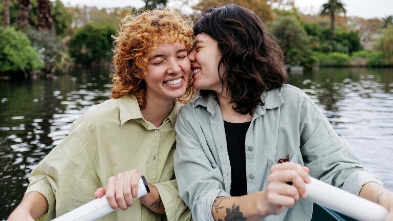 Zwei Frauen sitzen zusammen in einem kleinen Boot auf einem See. Eine Frau gibt der anderen einen inningen Kuss auf die Wange. Beide lächeln.