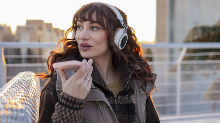 Eine Frau sitzt draußen auf einer Bank, trägt Bluetooth-Kopfhörer und hält ihr Handy vor den Mund.