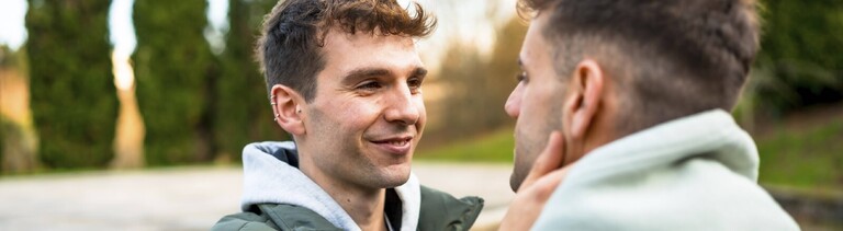 Zwei Männer stehen sich draußen gegenüber. Der eine Mann legt seine Hand an die Wange des anderen Mannes und lächelt. Beide tragen Winterjacken.