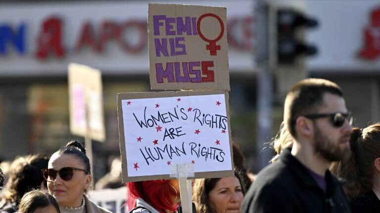 Auf einer Demo zum Weltfrauentag hält eine Teilnehmerin zwei Schilder in die Höhe, auf dem unteren steht "Women’s rights are human rights", auf dem oberen Feminismus mit zwei s (Frankfurt, 08.03.2025)
