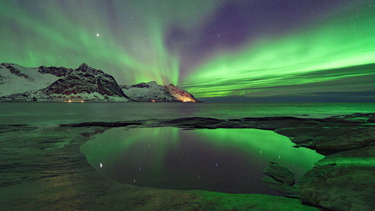 Der Nachthimmel über einer norwegischen Bucht schimmert grünlich. Am Horizont leuchten die Lichter einer Siedlung am Fuß eines Berges.