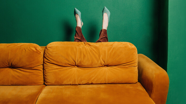 Die Beine einer Frau, mit grünen Absatzschuhen und einer braunen Hose, ragen hinter einem orangefarbenen Sofa hervor, die Wand dahinter ist grün