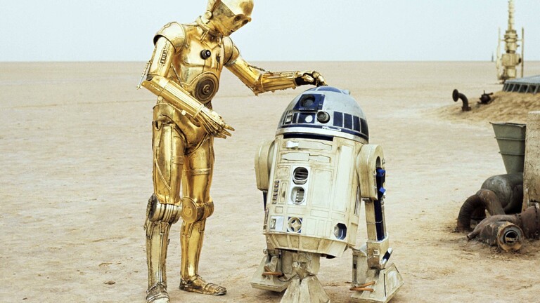Das Bild zeigt einen Ausschnitt aus der Filmreihe Star Wars. Zu sehen sind die beiden Charaktere C-3PO und R2-D2 in einer Wüstenlandschaft.