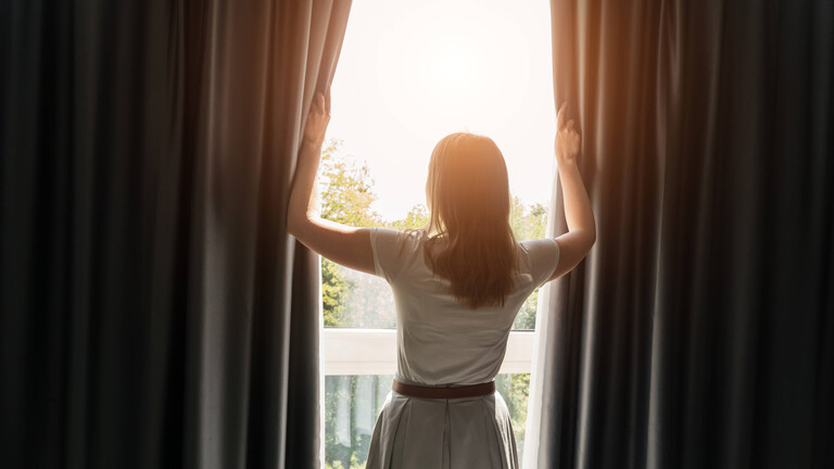 Frau öffnet Vorhänge in einem Zimmer am sonnigen Morgen.