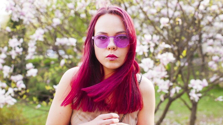 Eine Frau trägt eine rosafarbene Brille und steht im Freien vor einem rosablühenden Baum.