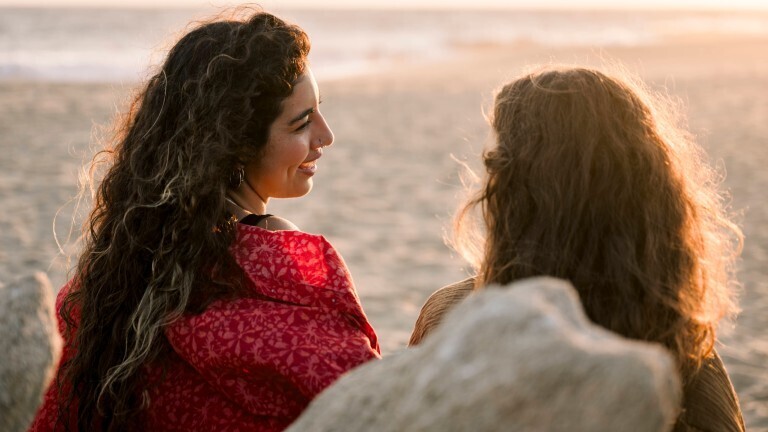 Zwei Frauen sitzen am Strand und unterhalten sich. Wir sehen sie von hinten.