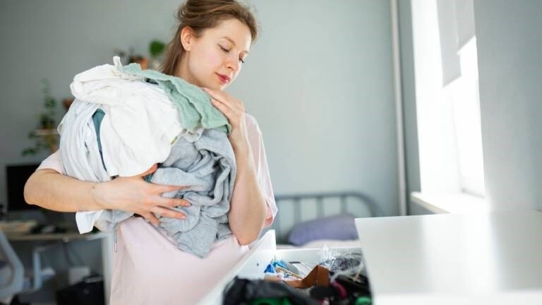 Eine Frau hat viele Wäsche auf dem Arm und steht vor einer Kommode mit geöffneter Schublade