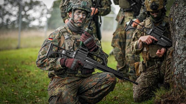 Vier Bundeswehrsoldaten tragen bei einer Übung Unform, Waffen, haben ihre Gesichter mit Tarnfarbe geschminkt und Stellung an einem Baum auf einer Wiese bezogen.