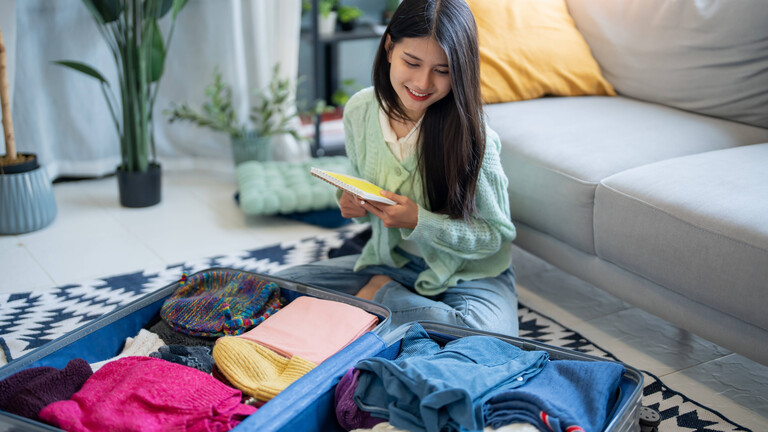 Foto einer jungen Frau, die lächelnd mit einer Packliste in der Hand vor einem bereits befüllten offenen Koffer sitzt