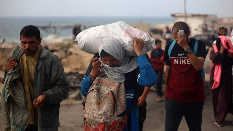 Frau im Gazastreifen trägt Hilfspaket auf dem Rücken und einen Rucksack auf ihrem Bauch (22.6.2025)