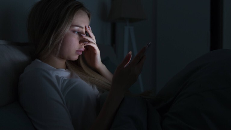 Eine junge Frau liegt in einem dunklen Zimmer in einem Bett. Das Licht ihres Handydisplays erhellt ihr Gesicht. Sie hält sich eine Hand vor das Gesicht.