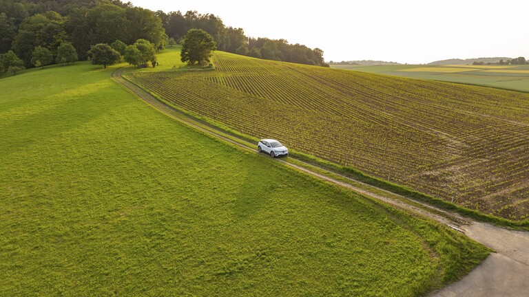 Ein E-Auto fährt auf einem Feldweg durch eine Wiesenlandschaft. Im Bild sieht man das E-Auto von oben.