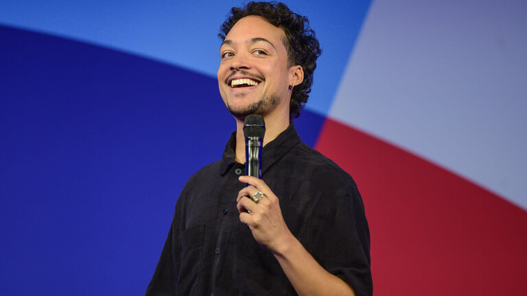Comedian und Moderator Aurel Mertz steht bei einer Veranstaltung auf der Bühne und hält ein Mikrofon in der Hand.
