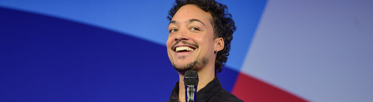 Comedian und Moderator Aurel Mertz steht bei einer Veranstaltung auf der Bühne und hält ein Mikrofon in der Hand.