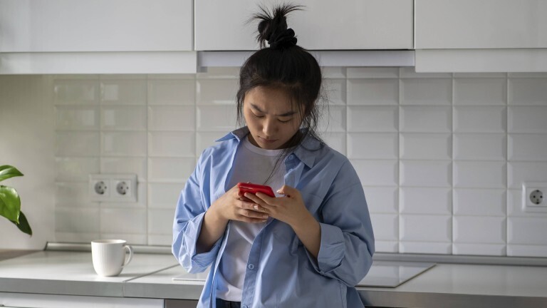 Eine junge Frau steht in einer Küche. Sie lehnt an der Küchenzeile an und schaut auf ihr Handy. Sie wirkt verärgert.