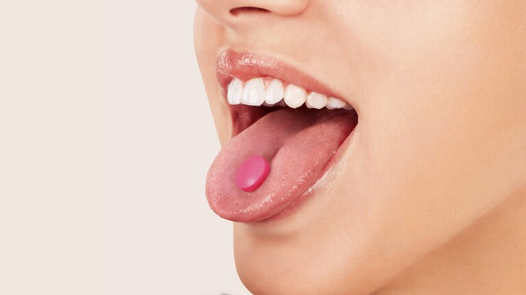 Eine Frau hat eine rosafarbene Tablette auf der Zunge, die sie rausstreckt.